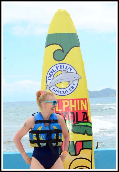 Sarah wearing swim suit and life jscket, stood in front of a large surf board promoting the dolphins
