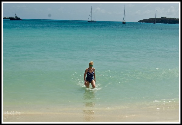 landscape shot of Sarah walking out of the turquoise sea, onto yellow sands, with sailing ships in the far distance