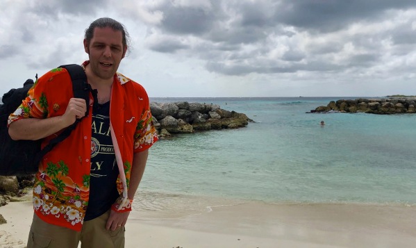Me stood on the left in a orange caribbean shirt carrying a black backpack with views of palm island, Aruba in the background