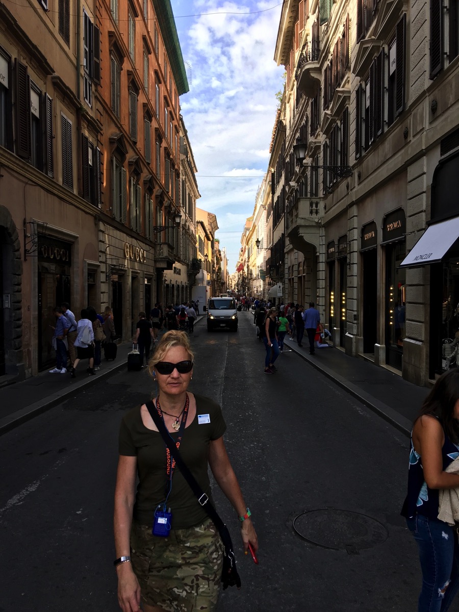 Sarah walking towards the camera down the centre of a busy street in Rome with tall building s on either side
